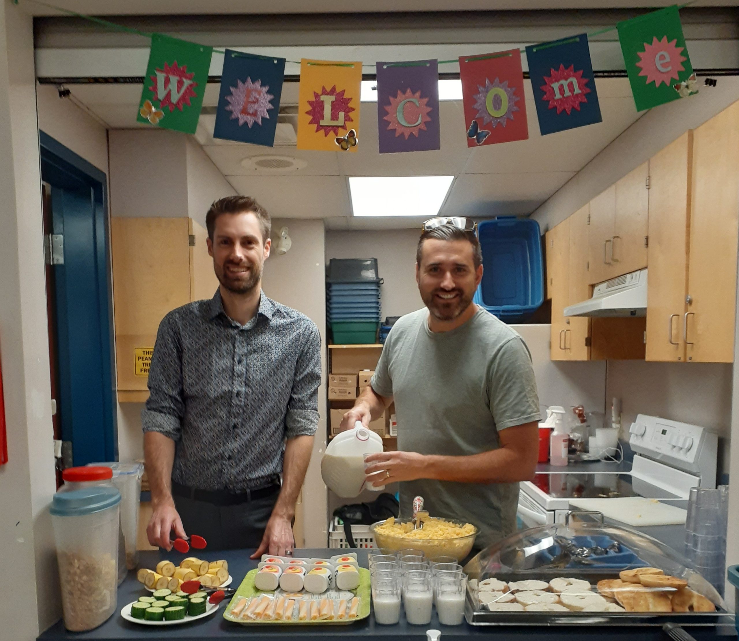 Two volunteers preparing a healthy breakfast station with milk, fruit, cereal, and snacks under a colorful Welcome banner for the school meal program.